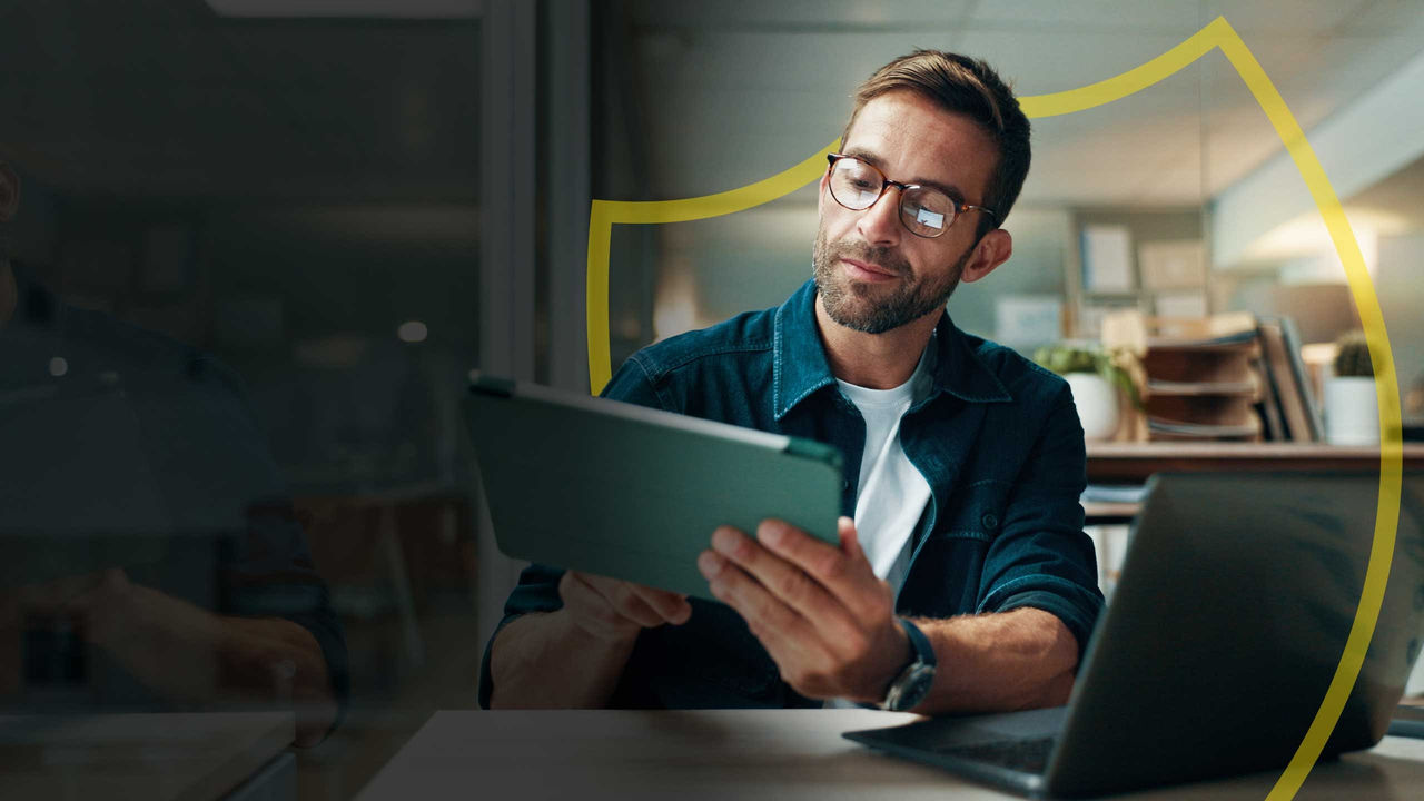 Business owener reviews transactions on his tablet with his laptop in the foreground.